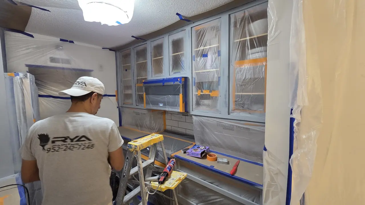 Painter masking kitchen cabinets with plastic sheeting and blue tape before refinishing