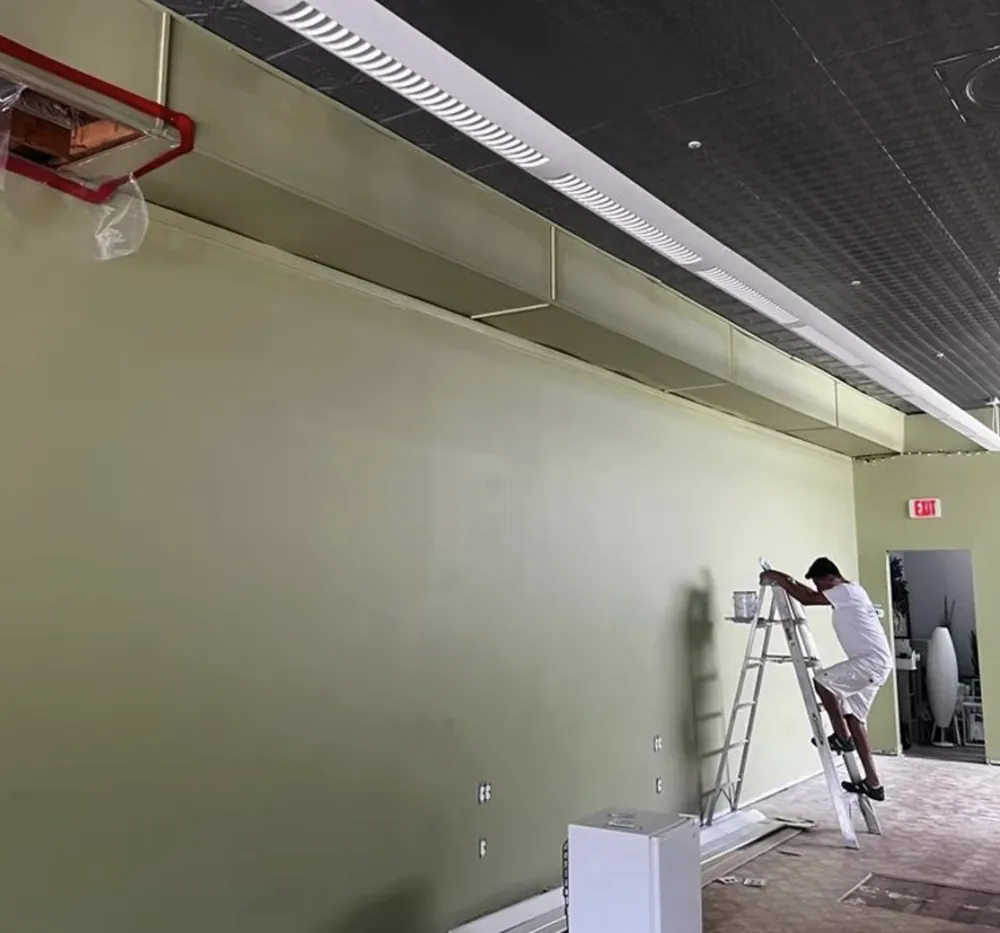 Painter on a ladder finishing an olive green wall in a commercial interior with black ceiling grid