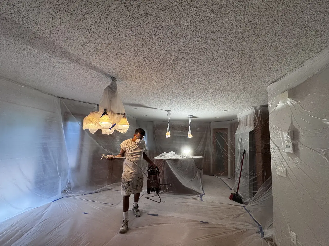 Painter in a respirator mask using a paint sprayer in a room fully covered in plastic sheeting