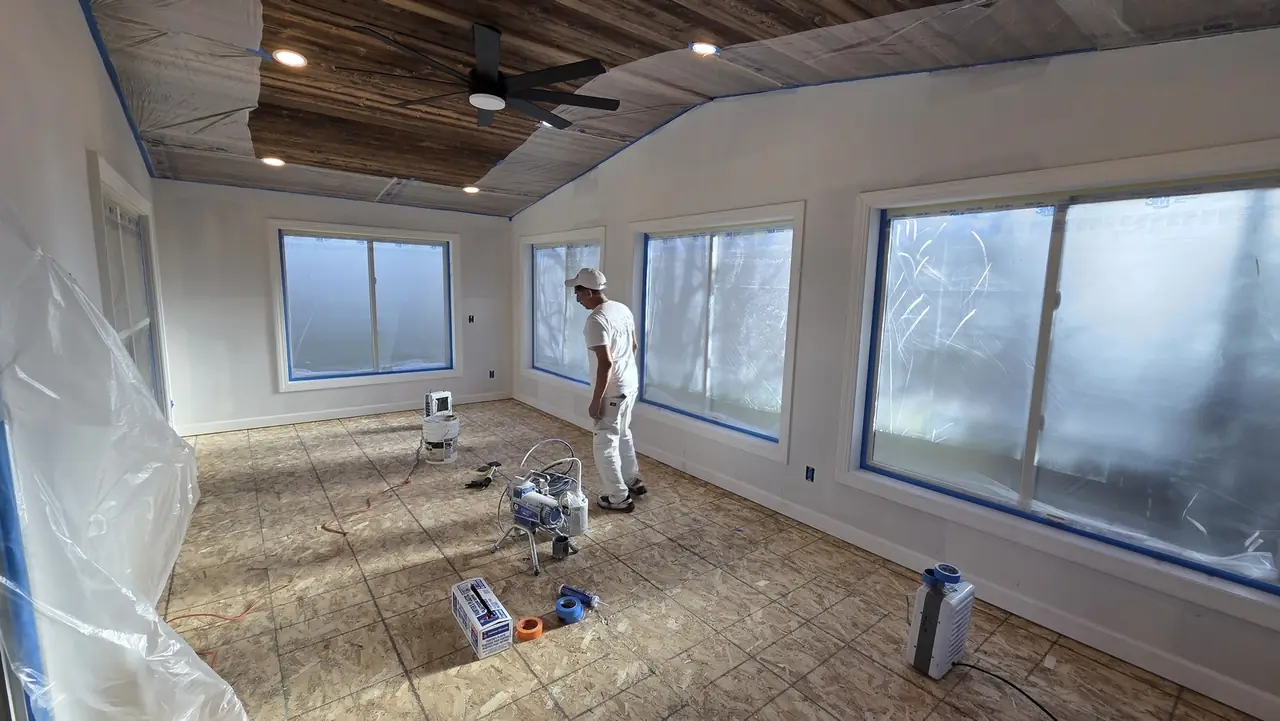 Painter using a paint sprayer in a large room with a vaulted wood ceiling and windows masked in plastic